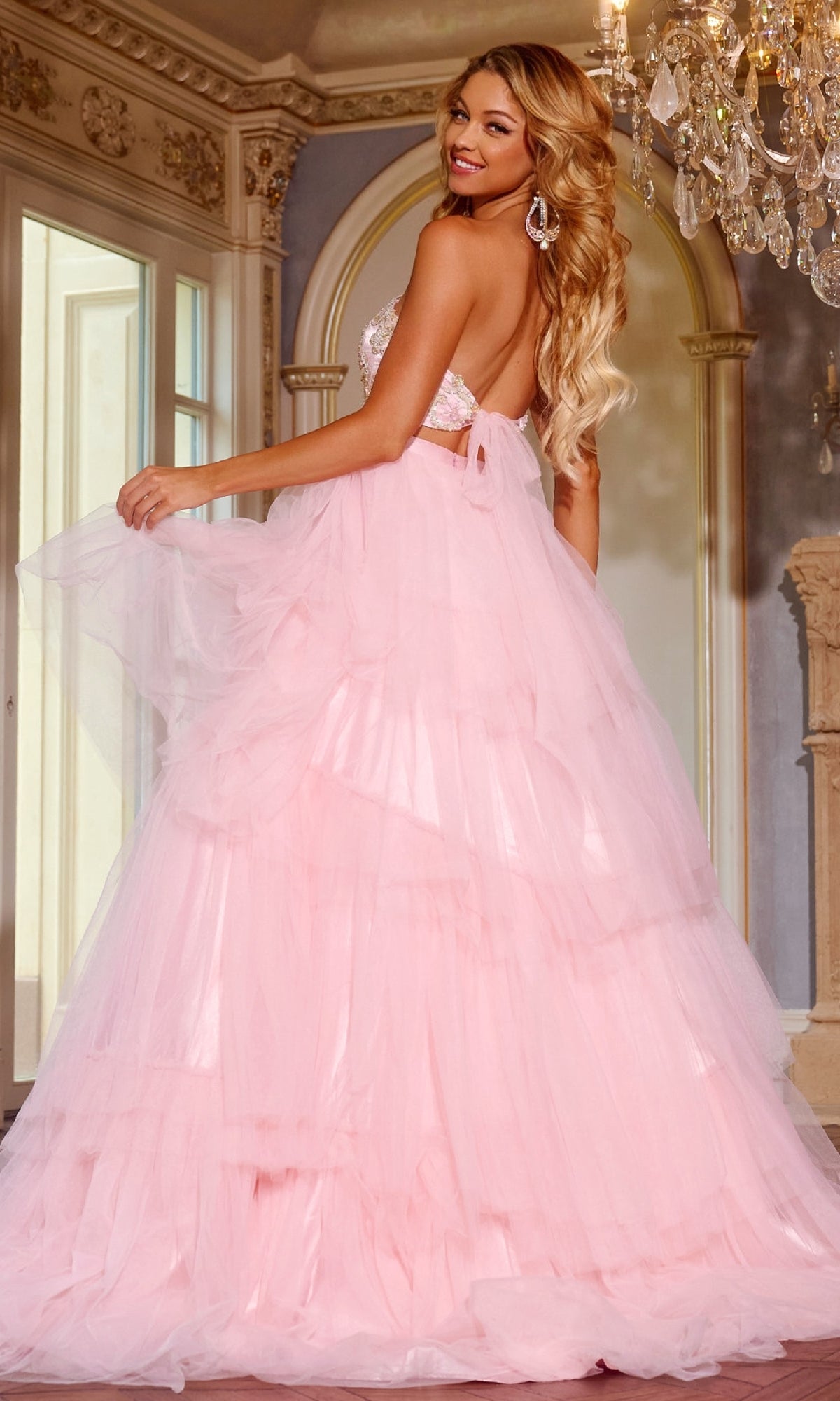 A woman in a light pink, two-piece Jovani D5112 prom ball gown with a floor-length ruffled tulle skirt stands in an elegant room, smiling over her shoulder.